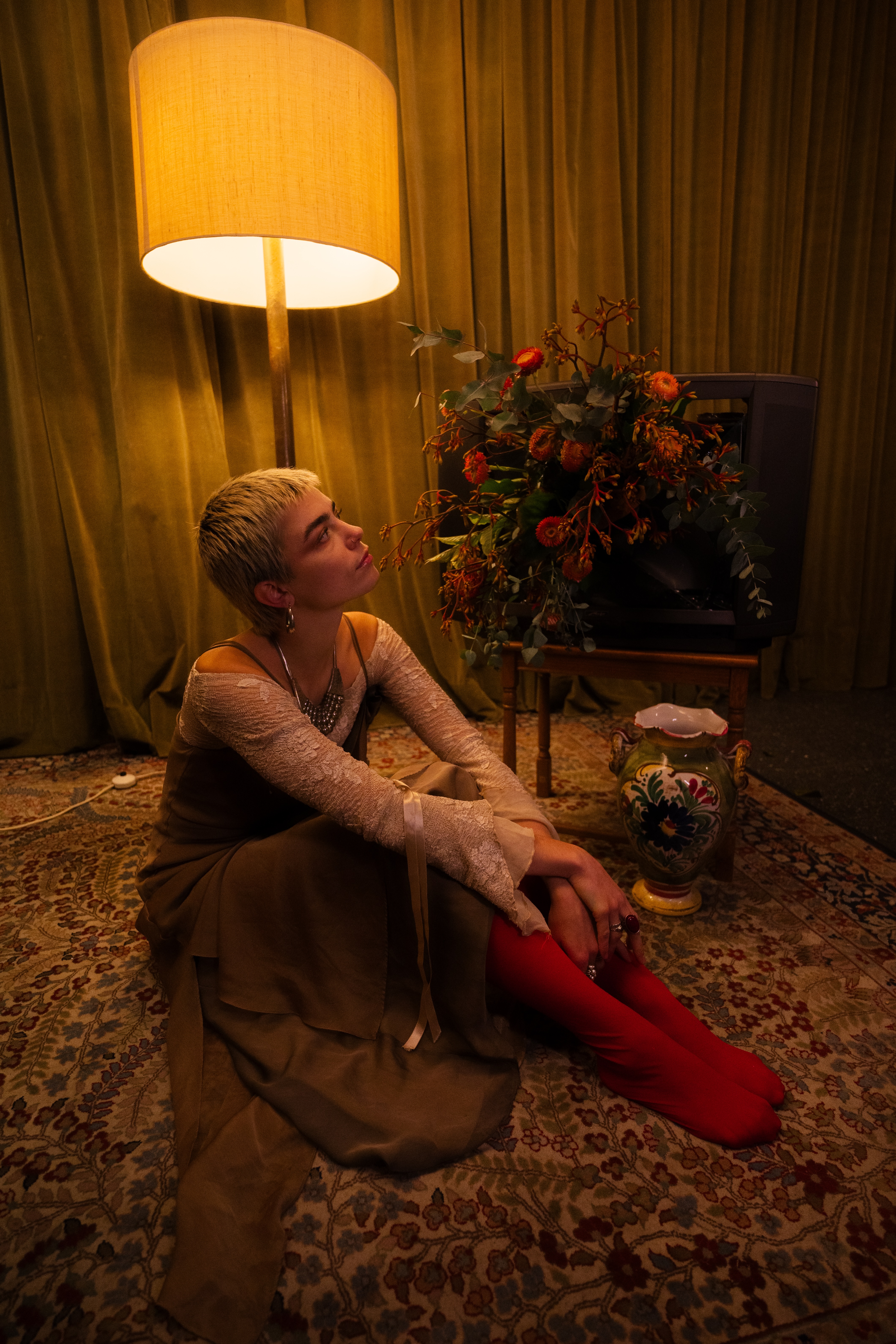 a woman with short hair, wearing a dress while sitting on the ground next to a dim lamp, an old tv and a vase